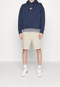 Felpa con cappuccio navy con logo, indossata sopra una camicia a righe, abbinata a pantaloni beige. Le scarpe sono bianche con accenti neri.