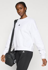 Λευκό φούτερ με λαιμόκοψη, που φέρει ένα μικρό μαύρο λογότυπο. Συνδυάζεται με μαύρο κολάν και μαύρη τσάντα με λευκές λεπτομέρειες.