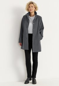 Frau in einem grauen Knopfmantel, weißem Hemd, schwarzen Hosen und schwarzen Stiefeln, die vor einem schlichten weißen Hintergrund steht.