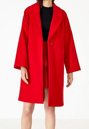 Kobieta ubrana w długi, jaskrawoczerwony płaszcz zapinany na jeden guzik, na czarnej bluzce i czerwonej spódnicy, sfotografowana na prostym, białym tle.