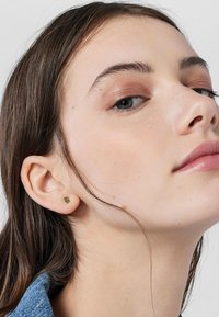 Pendientes de oro con una pequeña gema verde. Vista cercana del pendiente en la oreja de un modelo, mostrando detalles sutiles.
