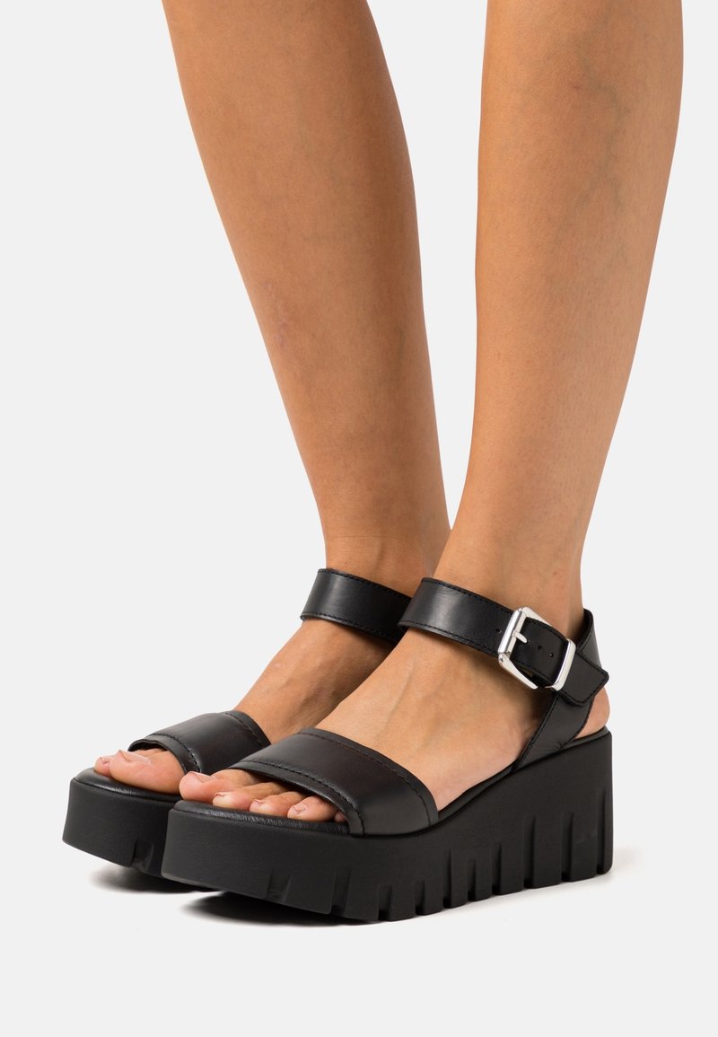 Sandales à plateforme noires avec semelles épaisses et brides à la cheville maintenues par des boucles argentées, portées sur des pieds et des jambes dénudés.