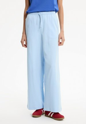 Persona que lleva pantalones de pierna ancha azul claro, una camiseta azul y zapatillas rojas con rayas blancas, de pie sobre un fondo liso.