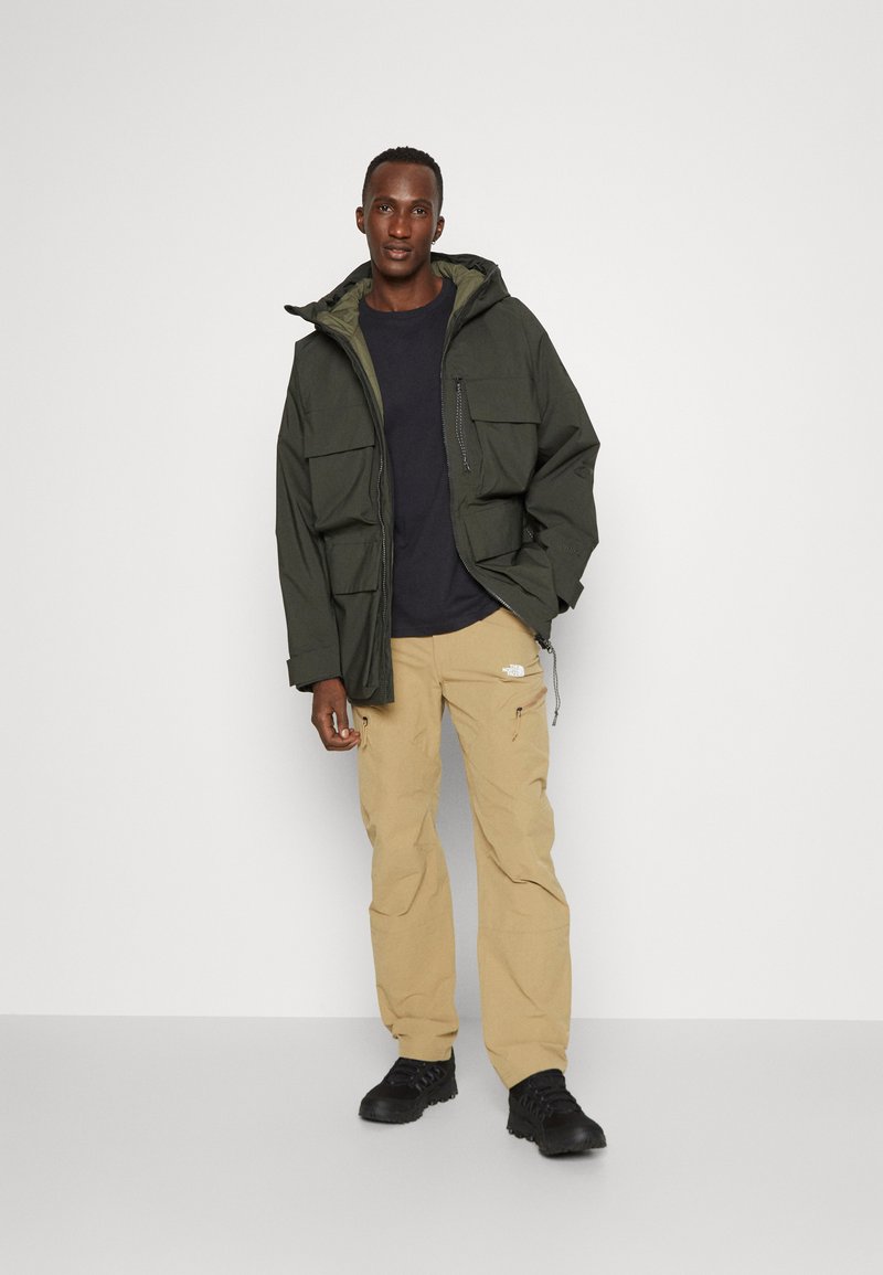 Chaqueta parka verde con grandes bolsillos frontales, llevada sobre una camiseta negra, combinada con pantalones cargo beige y zapatos negros. Diseño simple y funcional.