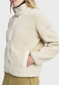 Chaqueta de forro polar en color crema, con cuello alto, cierre de cremallera, bolsillos laterales y un logo en la manga. Tela suave y texturizada.