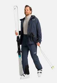 Attrezzatura da sci con una giacca impermeabile blu scuro, uno strato base grigio, pantaloni blu con dettagli neri, scarponi da sci bianchi e sci bianchi.