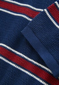 Texturerat marinblått tyg med röda och vita horisontella ränder. Har ribbade ärmslut och ett ventilerande vävt mönster.
