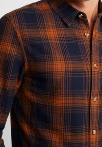 Camisa a cuadros en azul marino y naranja, con cuello abotonado, un patrón de cuadros distintivo y una textura suave. Botones de latón adornan el frente.