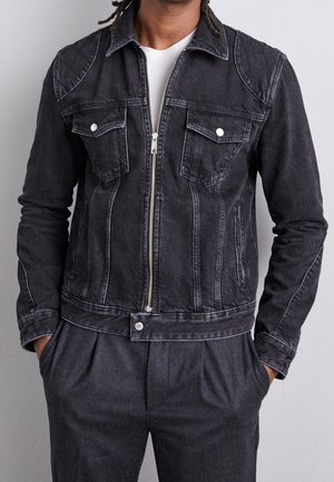 Giubbotto in denim nero con zip frontale, due tasche sul petto e colletto a punta. Presenta cuciture bianche, tasche laterali e un design aderente.