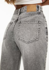 Jean en denim gris à taille haute, avec de grandes poches arrière, des coutures visibles et une étiquette de logo en cuir marron clair sur la ceinture.