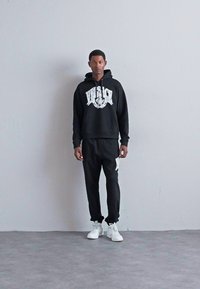 Uomo in piedi contro un muro neutro, indossa un hoodie nero di Versace, pantaloni della tuta neri abbinati con logo bianco e sneakers bianche.