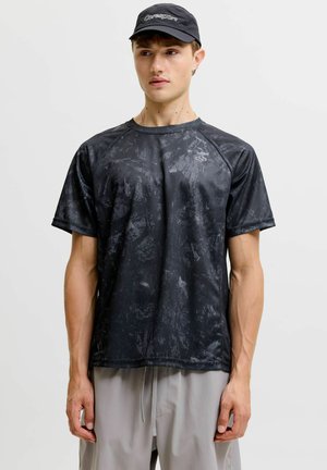 Jeune homme portant un t-shirt de sport noir à motif, un short gris clair et une casquette noire avec le logo "Coresport", debout devant un fond uni.