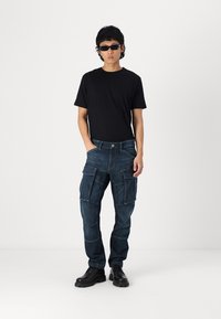 Dark denim cargo pants with multiple pockets, fitted black t-shirt, and black sunglasses. Model stands in a neutral setting.