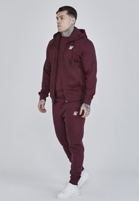 Bordeauxrode zip-up hoodie en trainingsbroek set gemaakt van zachte stof. Kenmerkend is het geborduurde logo in het wit. Gecombineerd met witte sneakers.