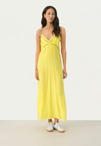 Gelbes Maxikleid mit Spaghettiträgern, das über einen V-förmigen Ausschnitt und eine mittige Verzierung verfügt. Das Model kombiniert es mit weißen Sneakers.