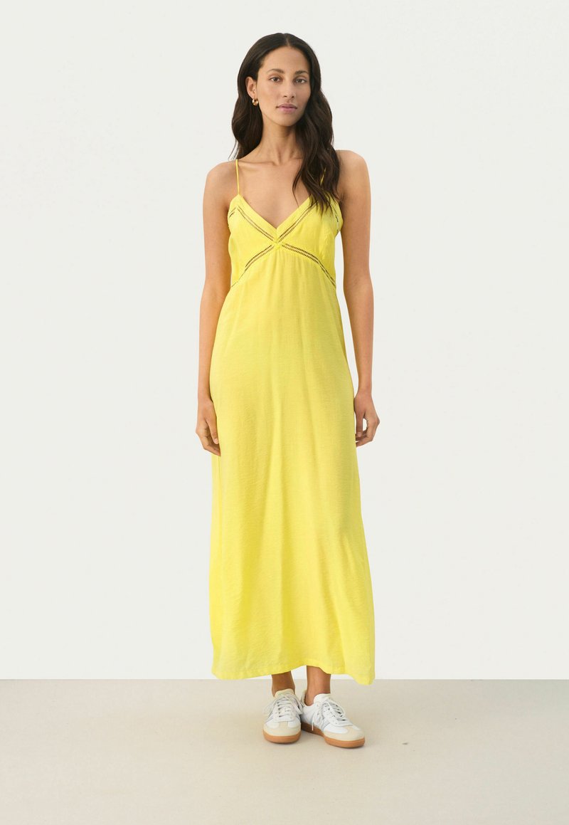 Gelbes Maxikleid mit Spaghettiträgern, das über einen V-förmigen Ausschnitt und eine mittige Verzierung verfügt. Das Model kombiniert es mit weißen Sneakers.