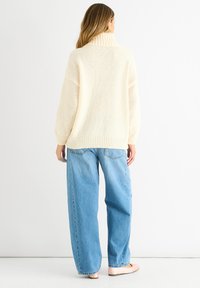 Maglione oversize beige chiaro in maglia con colletto alto a coste, abbinato a jeans di denim blu larghi. Texture morbida e vestibilità rilassata.