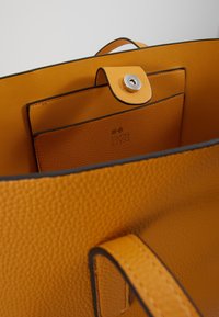 Borsa a tracolla in pelle arancione con texture, dotata di una tasca interna chiusa da un bottone a scatto magnetico e di un piccolo manico visibile all'interno.