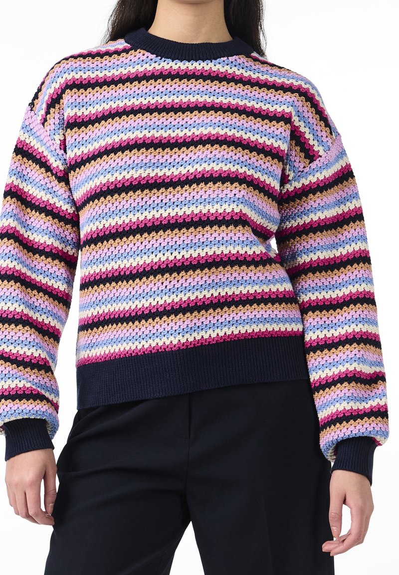 Femme portant un pull en tricot avec des rayures horizontales bleu marine, rose, violet, beige et bleu clair, assorti à un pantalon noir.
