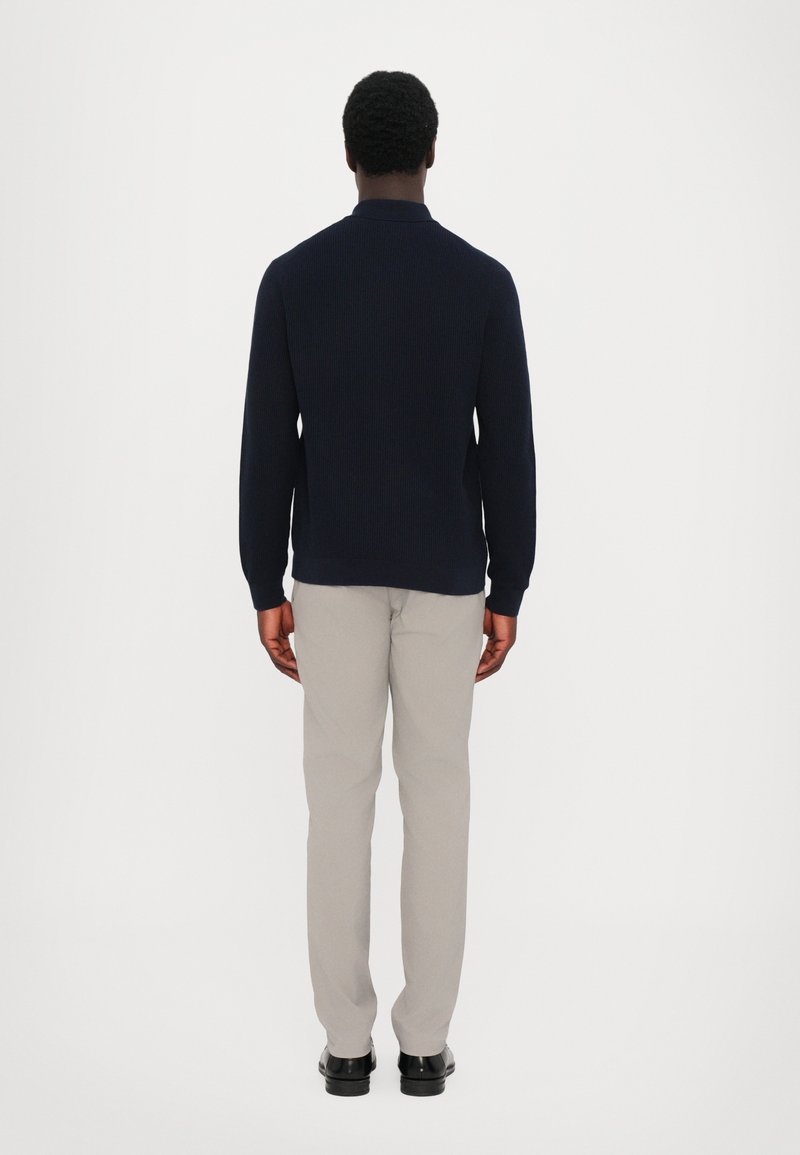 Marineblå strikpullover med høj krave, parret med lysegrå bukser og sorte sko. Enkelt, rene linjer og subtil tekstur.