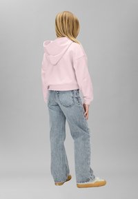 Sudadera corta de color rosa claro con capucha, combinada con vaqueros de color azul claro y corte holgado. El conjunto presenta una silueta relajada y un diseño sencillo.