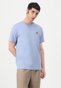 T-shirt en coton bleu clair avec un col rond, des manches courtes et un petit logo de papillon jaune sur la poitrine gauche. Texture douce.