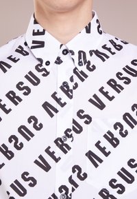 Witte blouse met een herhalend groot zwart "VERSUS" tekstpatroon. Zwarte knopen en een borstzakje maken het ontwerp af.