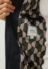 Marinejakke med et mønstret indre, der fremviser store grønne og beige cirkler, der skifter med striber, samt et mærke med teksten "NEXT TAILORING."