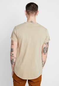 Beige t-shirt met korte mouwen en een ronde onderrand, gemaakt van zachte stof. Tatoeages zichtbaar op de armen. Model staat met de rug naar de camera, draagt een broek in bruin.