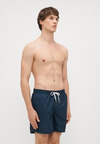 Tumed ujumispüksid, millel on peen vertikaalne triipude muster, elastne vöökoht ja valge pael, valmistatud kergest ja hingavast materjalist.