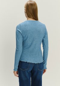 Donna con capelli lunghi, che indossa un maglione fitted azzurro chiaro con un motivo e jeans blu scuro, in piedi di spalle contro uno sfondo neutro.