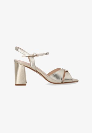 Metallische gouden blokhaksandalen met een slanke enkelband, open teen, kruislings ontwerp aan de voorkant en een gladde, glanzende textuur.
