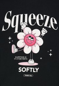 Schwarzes Shirt mit einer bunten Cartoon-Blume mit einem lächelnden Gesicht, umgeben von Sternen. Text: "Sanft drücken" und "Happiness ist in allem."