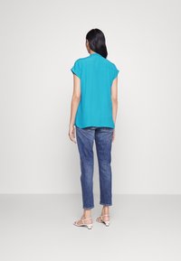 Camicia turchese ampia a maniche corte, vista posteriore, abbinata a jeans blu e sandali bianchi con cinturini su uno sfondo semplice.