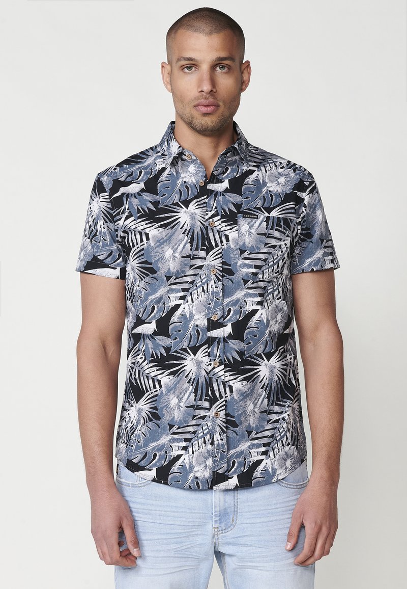 Camicia a maniche corte con bottoni, caratterizzata da uno sfondo nero e da un motivo di foglie tropicali blu e bianche. Include bottoni marroni e una tasca sul petto.