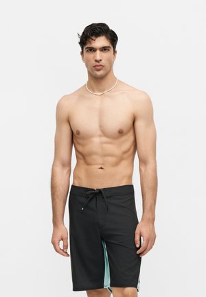 Młody mężczyzna z krótkimi ciemnymi włosami, ubrany w czarne spodenki kąpielowe i biały naszyjnik z muszli, stojący na jednolitym jasnym tle.