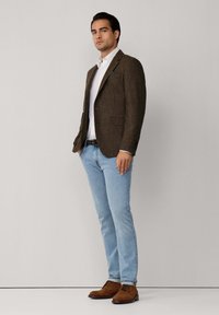 Muškarac u bijeloj košulji, smeškastom kariranom sako, svijetloplavim trapericama, smeškastim nubuk cipelama, stoji naslonjen na ravni svijetlu zid.