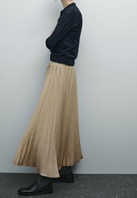 Hame pleatattu, beige väri, valmistettu teksturoidusta kankaasta, yhdistetty tumman sävyiseen neuleeseen ja mustiin loafer-kenkiin, esitetty profiilikuvassa yksinkertaista taustaa vasten.