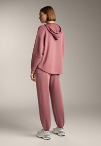 Ružová mikina s kapucňou a voľným strihom, v kombinácii s kožami s užším strihom. Mäkka tkanina, elastické manžety a výrazné detaily šitia.