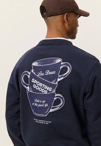 Μπλε ναυτικό φούτερ με γραφικό στοιχείο στοίχισης που απεικονίζει φλιτζάνια καφέ σε λευκό χρώμα με το κείμενο «Les Deux SPORTING GOODS» και «Απόλαυσε μια γουλιά καλής ζωής.»