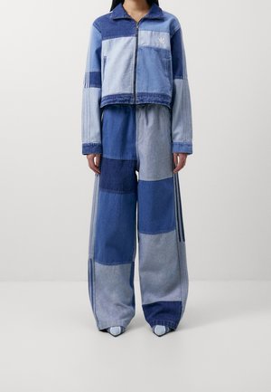 Person wearing a patchwork denim jacket and wide-leg pants in various shades of blue, standing against a plain white background.