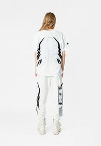 Camisa blanca de gran tamaño con bordado detallado en negro y dorado en la parte posterior representando alas. Combinada con pantalones de chándal blancos con bolsillo lateral y etiqueta.