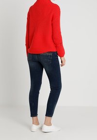 Czerwony sweter z wysokim kołnierzem, dziany, o prążkowanej fakturze, zestawiony z ciemnoniebieskimi, obcisłymi dżinsami i białymi butami typu slip-on.