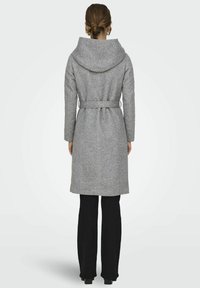Manteau en laine gris avec une grande capuche, taille ceinte et manches longues, arrivant au milieu du mollet. Associé à un pantalon noir et des bottes à talons.