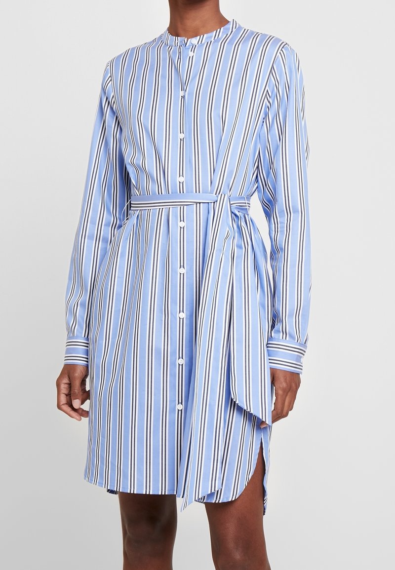 Femme portant une robe boutonnée à rayures verticales bleues et blanches, à manches longues et avec une ceinture nouée à la taille.