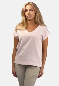 Jasnoróżowa bluzka z krótkim rękawem w dekolcie w kształcie litery V, gładka w dotyku i o prostym wykończeniu. Mały metalowy akcent z logo w pobliżu dołu.