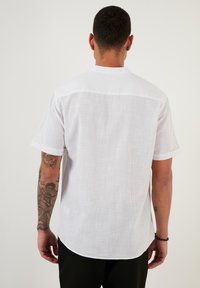 Cămașă albă din in cu mânecă scurtă, având un spate drept, guler tip pliants și o croială lejeră. Model texturat subtil vizibil în material.