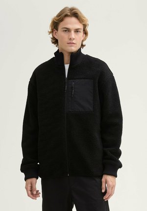 Schwarze Fleecejacke mit hohem Kragen und einer mit Reißverschluss versehenen Brusttasche. Hat eine strukturierte Außenseite, gerippte Bündchen und eine lockere Passform.