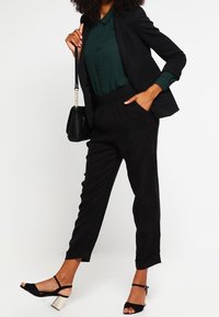 Zwarte op maat gemaakte broek met een elastische tailleband, gecombineerd met een donkergroene blouse en een zwarte blazer. Heeft strapschoenen met een chunky hak.