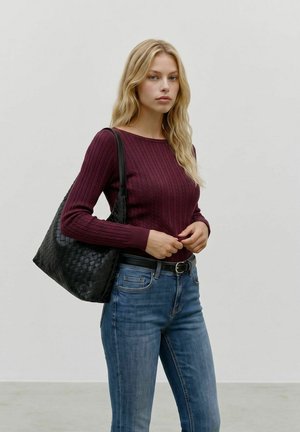 Jeune femme aux cheveux blonds porte un pull bordeaux et un jean bleu, portant un grand sac à bandoulière en cuir tressé noir.